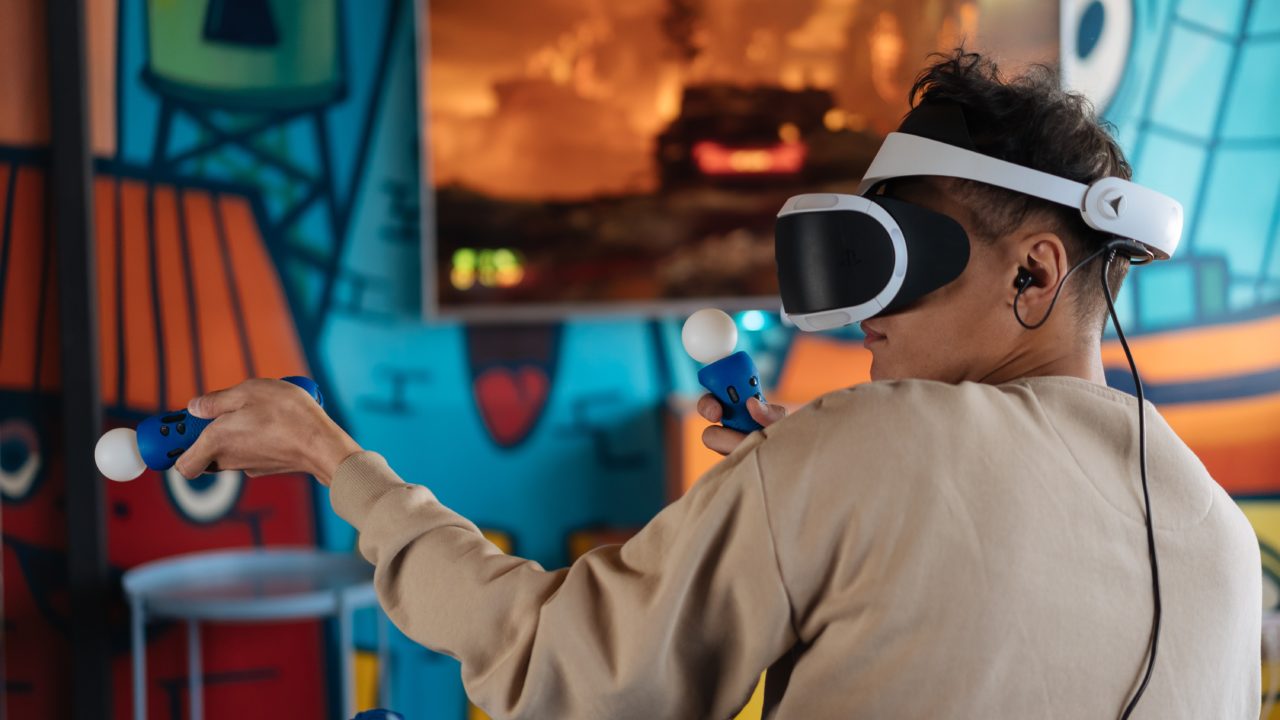 Un jeune homme, équipé d'un casque de réalité virtuelle, tient deux manettes dans ses mains sur un fond coloré.